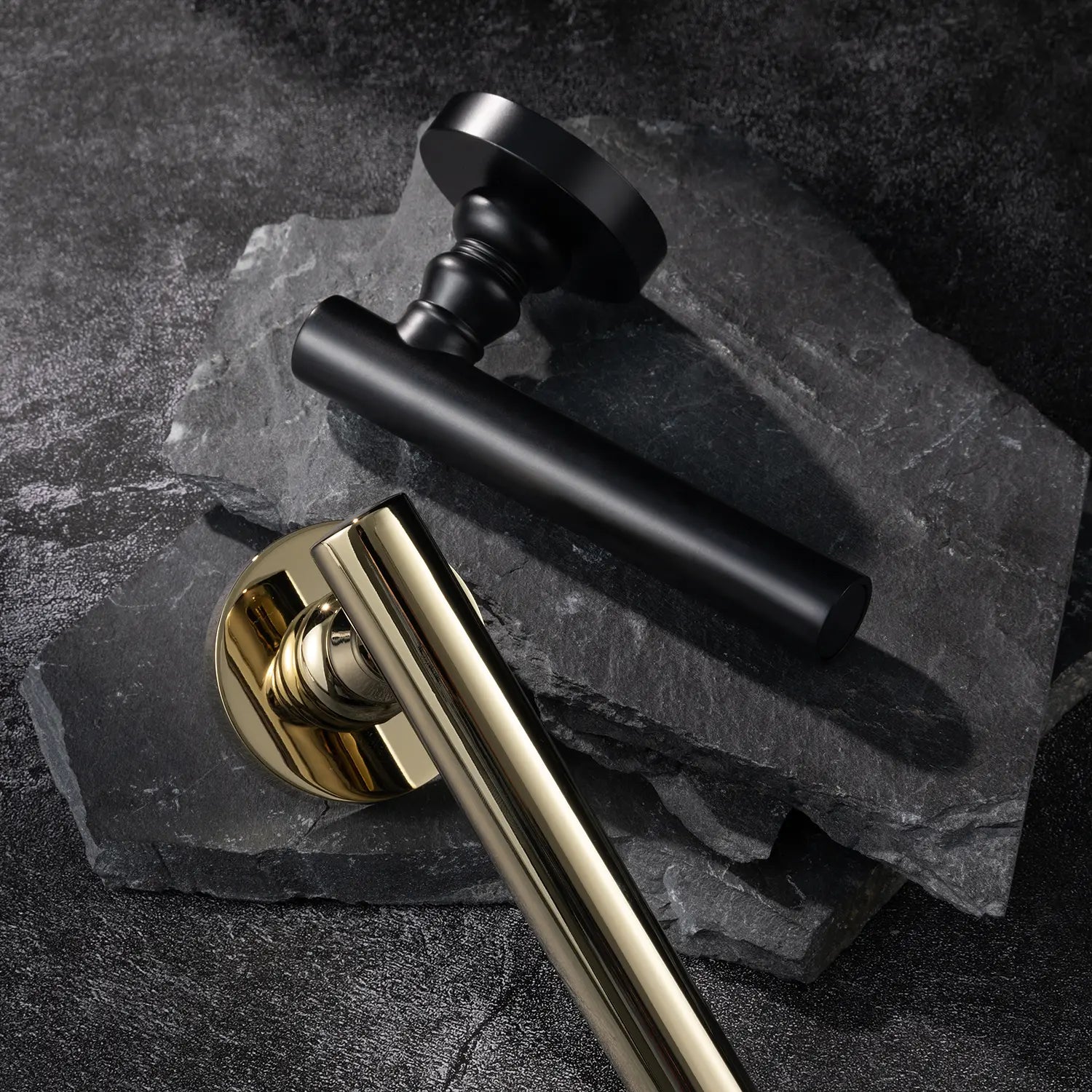 Two Alda door handles in polished brass and black finishes, displayed together on a textured stone background.