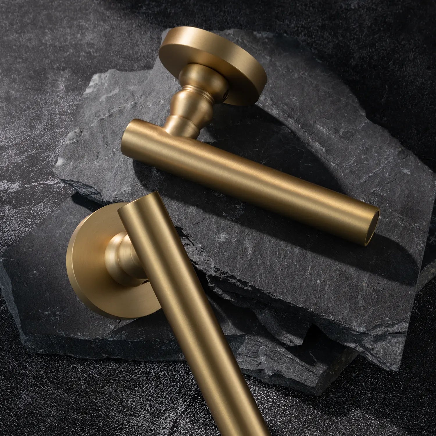 Two Alda door handles in satin brass finish, displayed on a textured stone background.