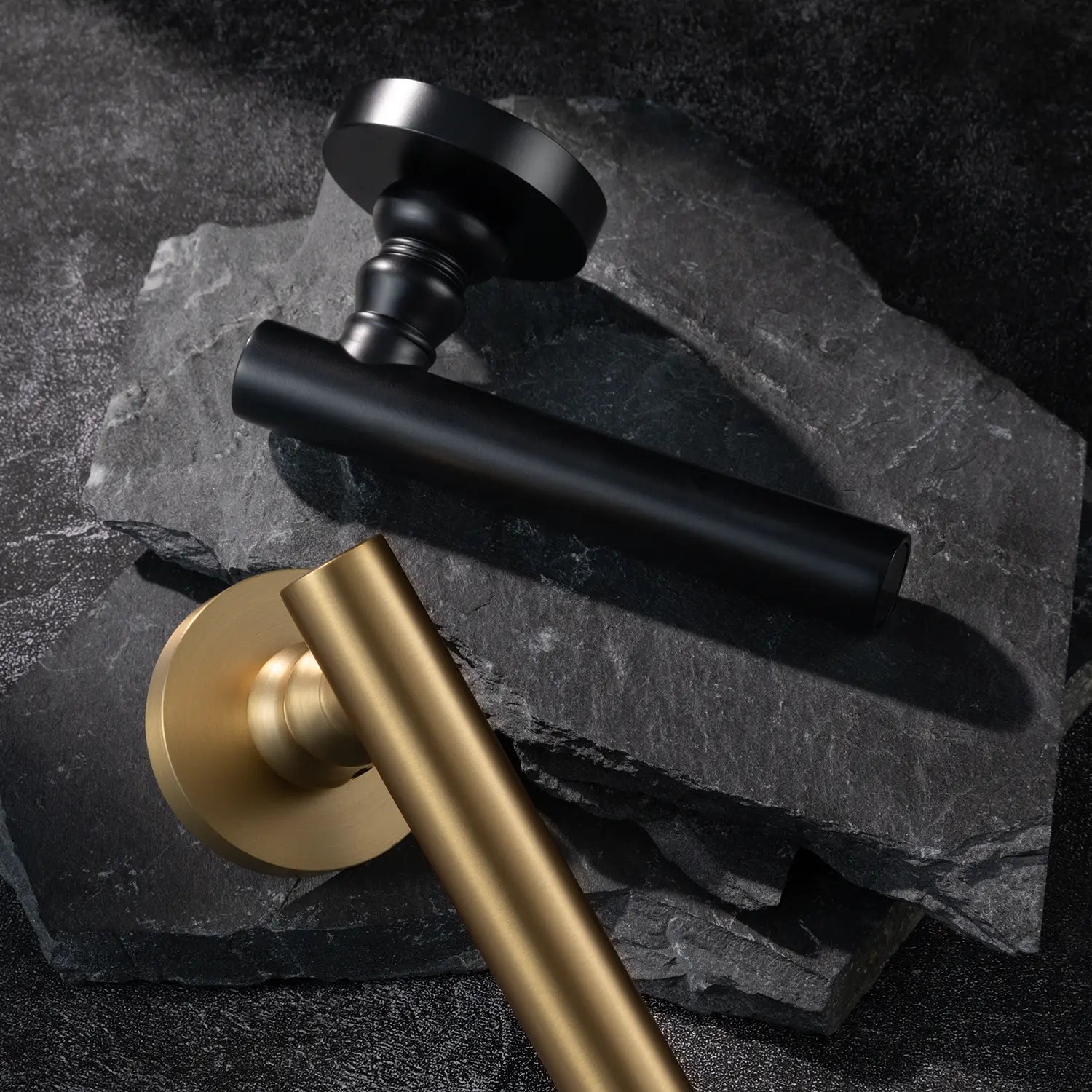Two Alda door handles in satin brass and black finishes, displayed together on a textured stone background.