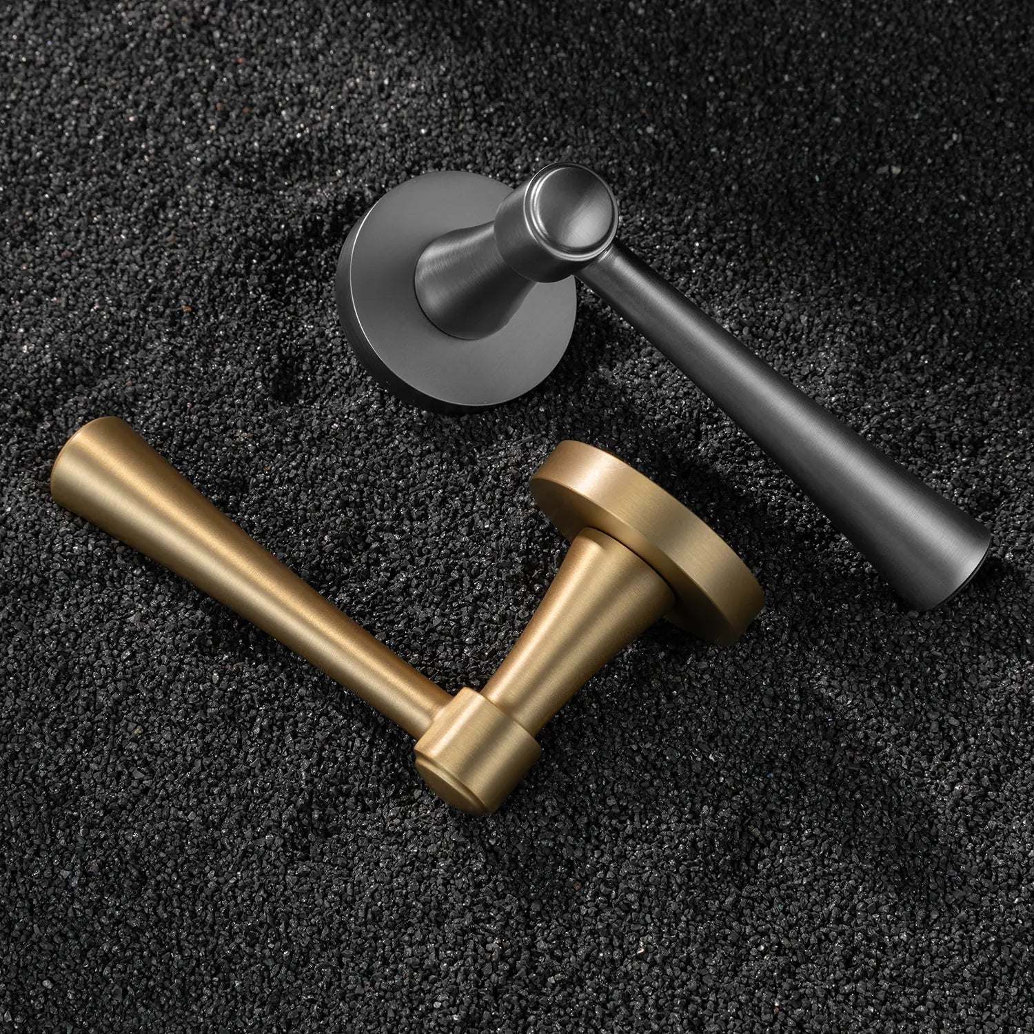 Two Hilda door handles in satin brass and charcoal grey finishes, elegantly positioned on a textured black surface. Featuring sleek and timeless cone-shaped design for modern interiors.