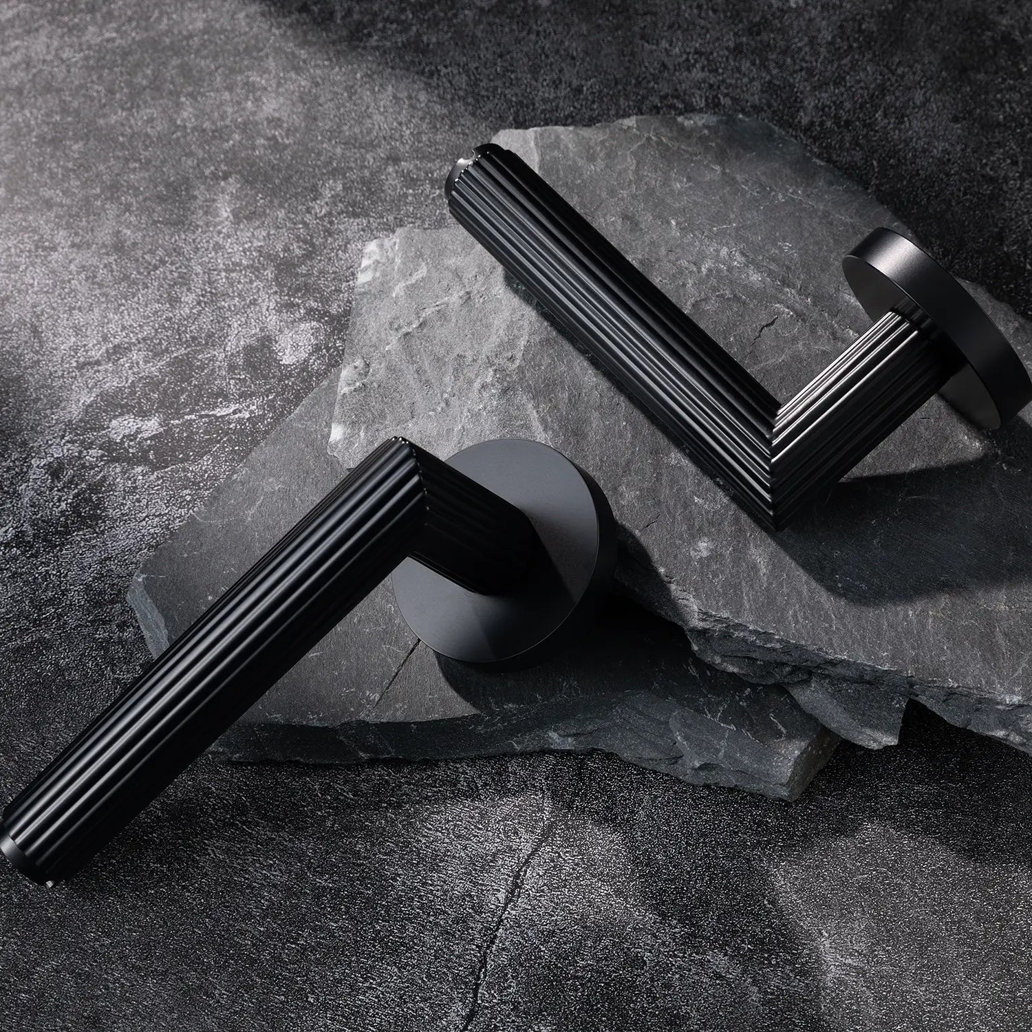 Two Midal door handles in black finish, displayed diagonally on a textured stone surface, featuring a bold linear pattern for a contemporary aesthetic.