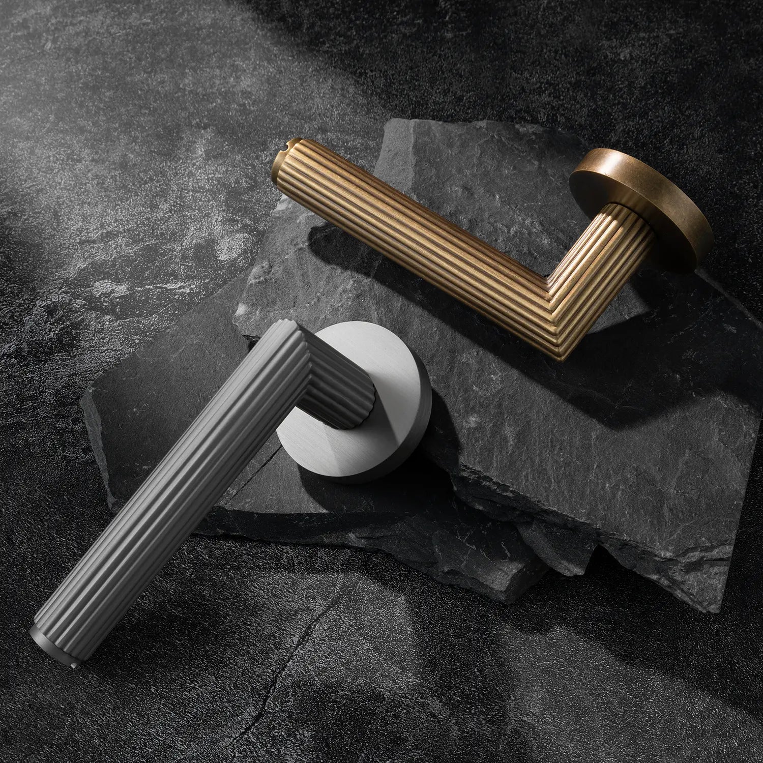 Two Midal door handles in nickel and ochre antique brass finishes, displayed diagonally on a textured stone surface, featuring a bold linear pattern for a contemporary aesthetic.