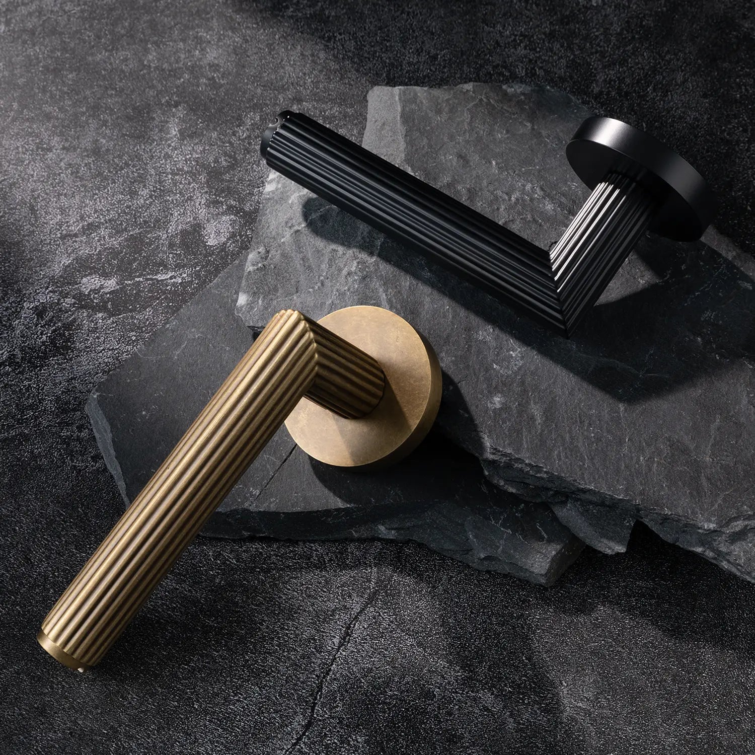 Two Midal door handles in black and antique brass finishes, displayed diagonally on a textured stone surface, featuring a bold linear pattern for a contemporary aesthetic.