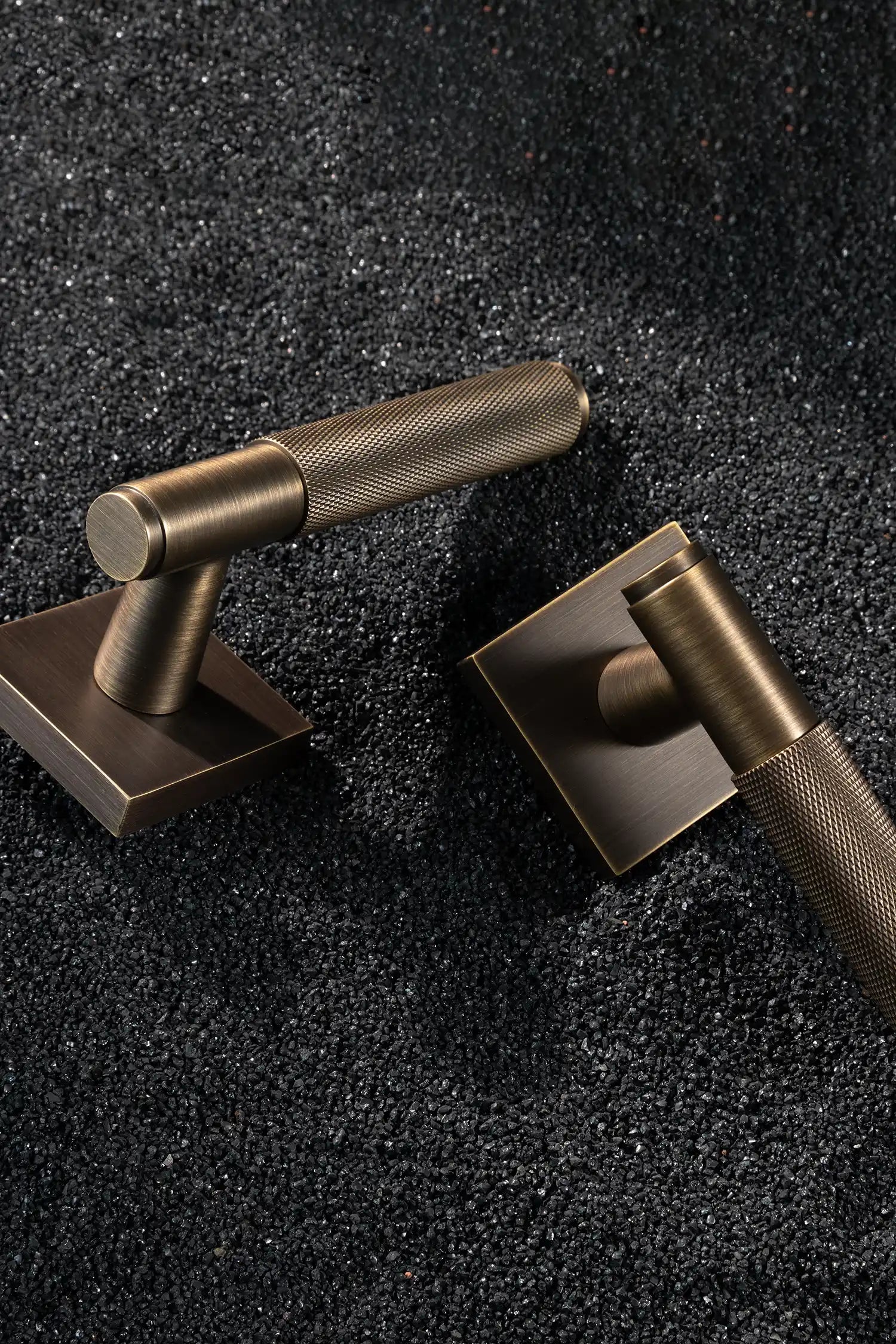 Svik-01 internal door handles in antique brass finish, featuring knurled pattern grips, paired with square rose bases, displayed on a textured black gravel background.