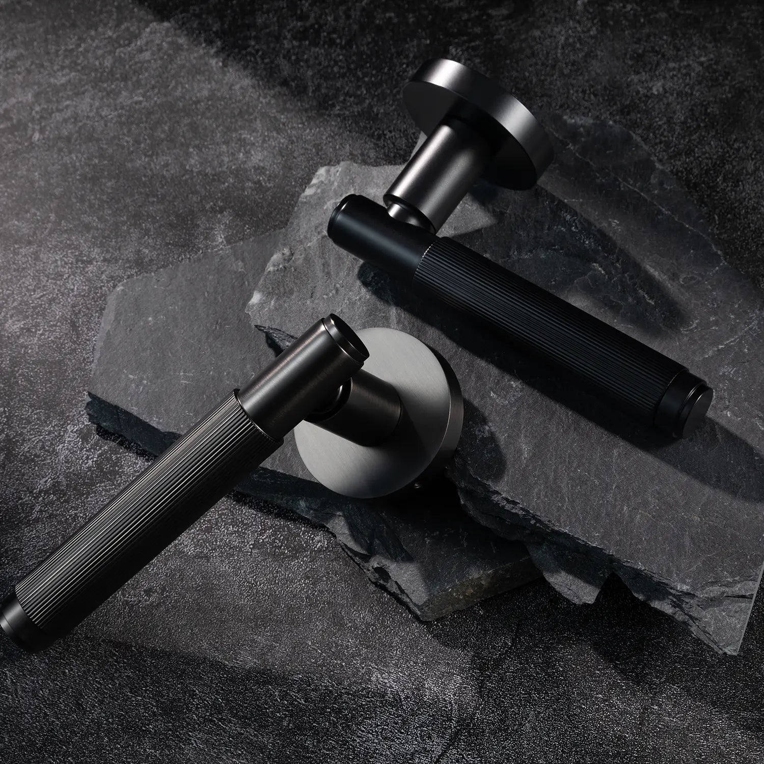 Two Svik-02 internal door handles in charcoal grey and black finishes, positioned on dark textured stone, highlighting contrasting finishes with linear pattern details.