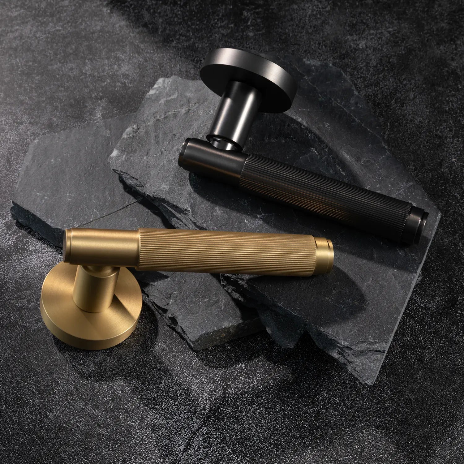 Two Svik-02 internal door handles in satin brass and charcoal grey finishes, positioned on dark textured stone, highlighting contrasting finishes with linear pattern details.