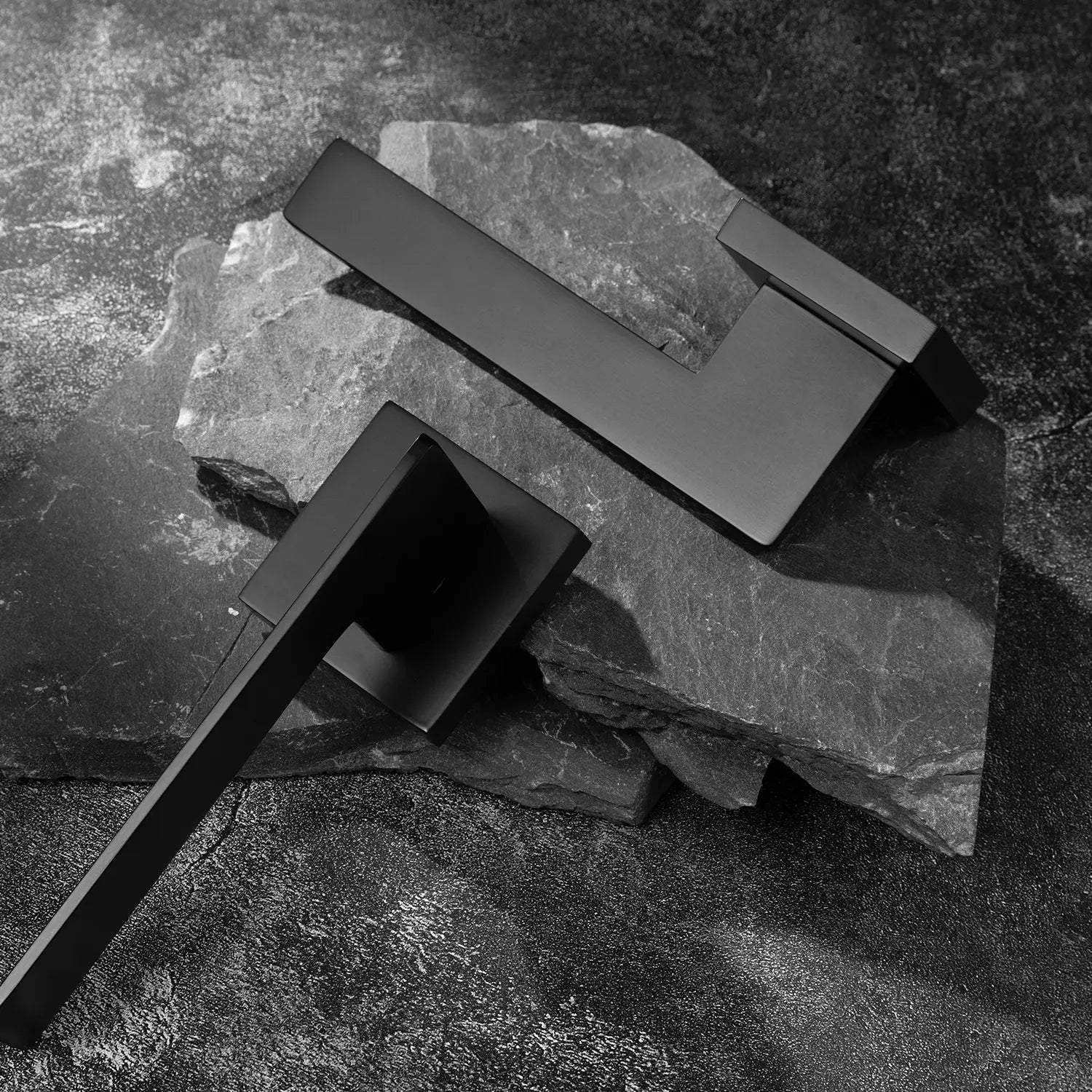 Two Tagg internal door handles in black finish, featuring a minimalist geometric lever design and square rose, displayed on two overlapping stone slabs.