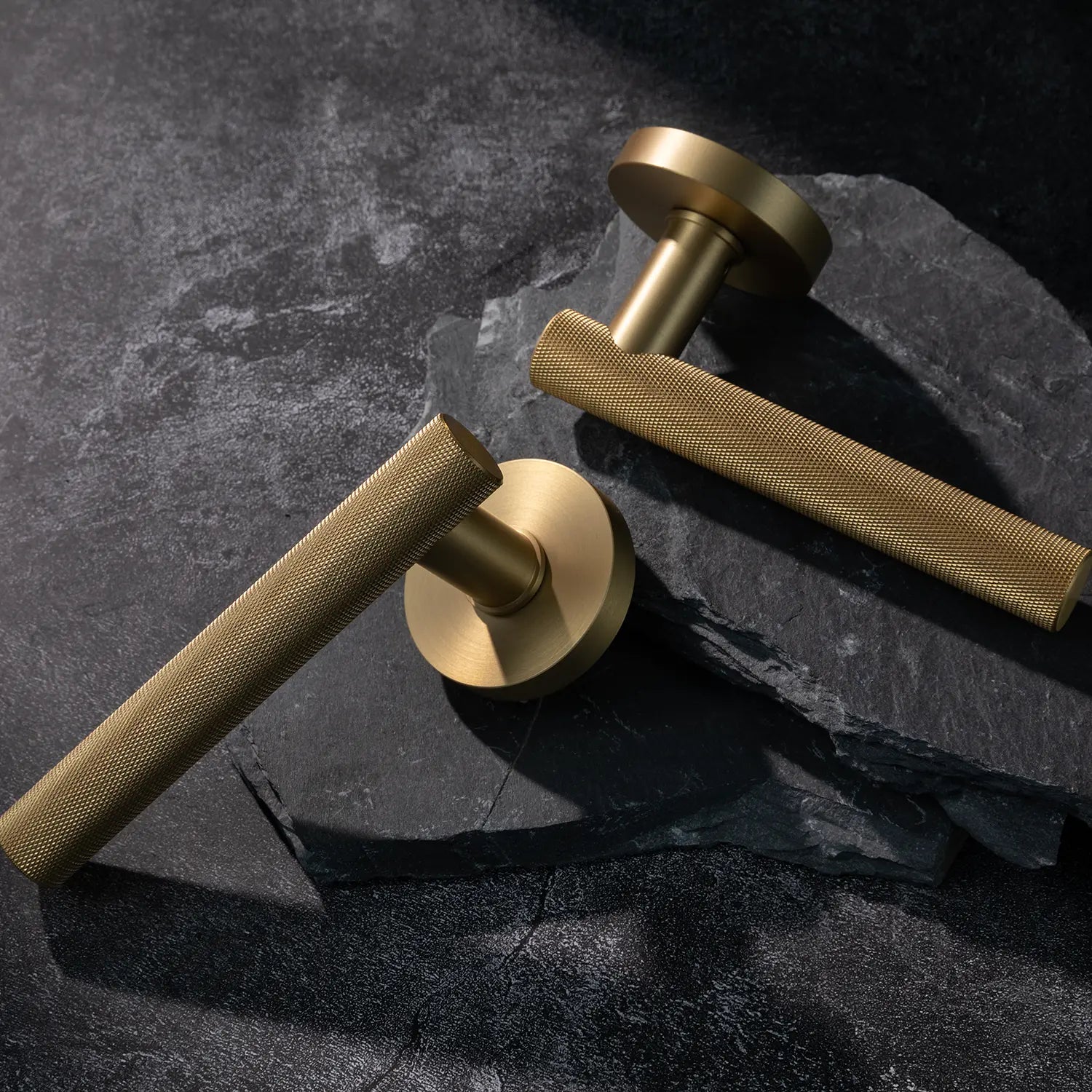 Two Tord-01 internal door handles in satin brass finish, featuring a fully knurled pattern handle design, placed diagonally on stacked stone slabs with a textured background.