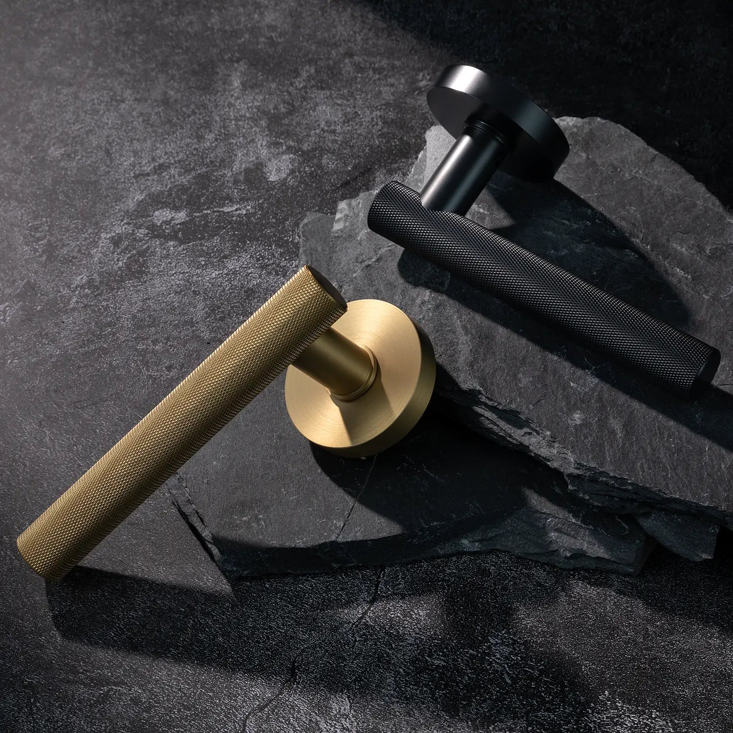 Two Tord-01 internal door handles, one in satin brass and the other in black finish, both featuring a fully knurled pattern handle design, placed diagonally on stacked stone slabs with a textured background.