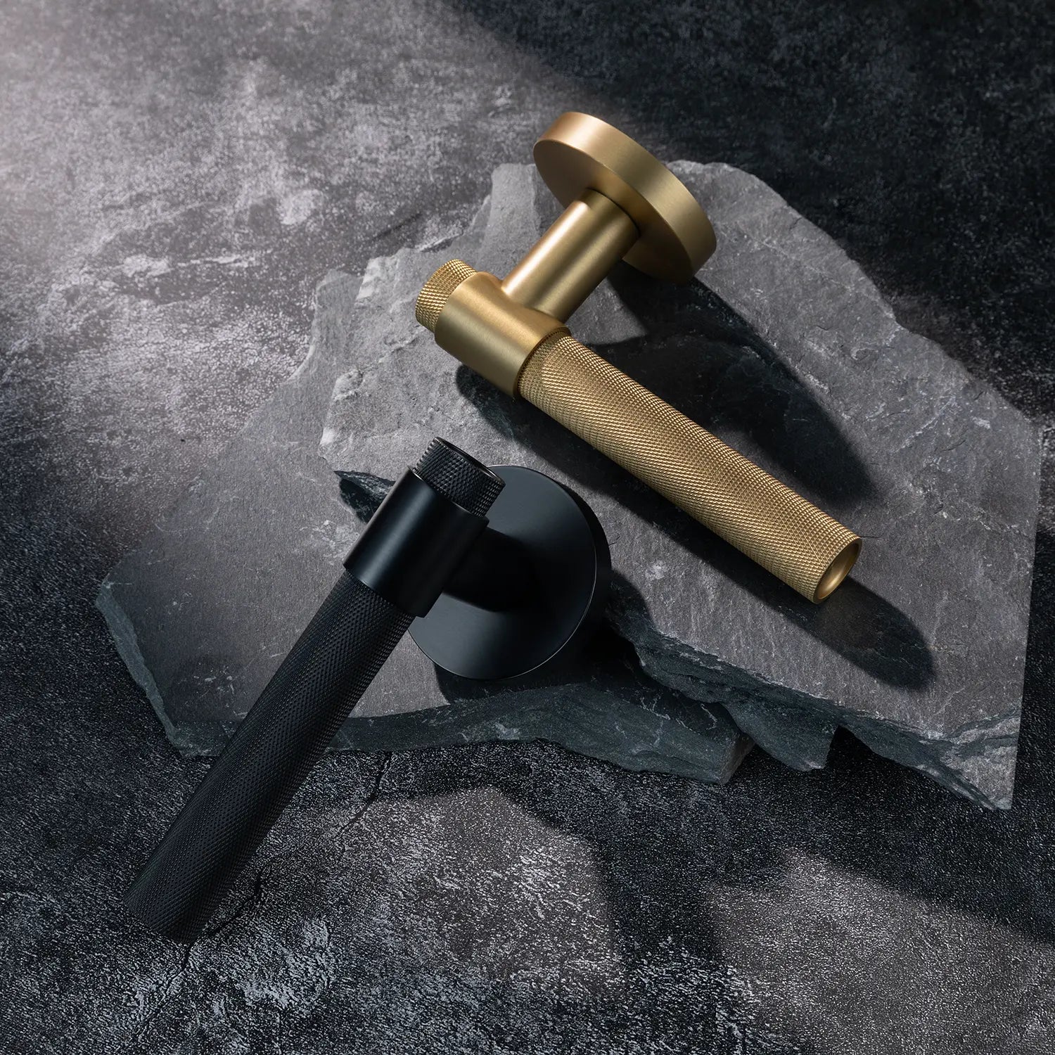 Two Tord-02 internal door handles in satin brass and black finishes, featuring large knurled pattern grips, positioned on overlapping dark stone slabs.