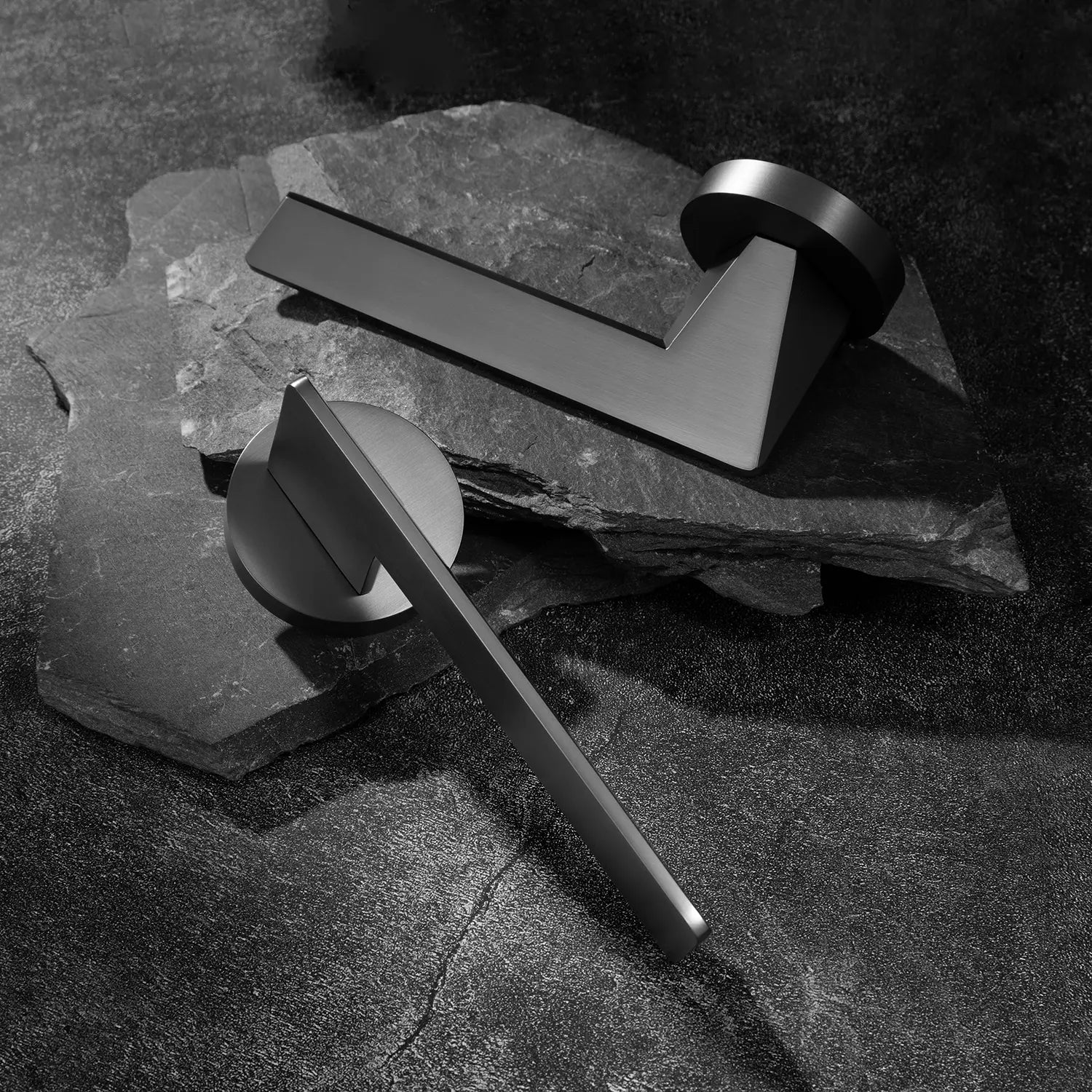 Two Tagg internal door handles in charcoal grey finish, featuring a minimalist geometric lever design and round rose, displayed on two overlapping stone slabs.