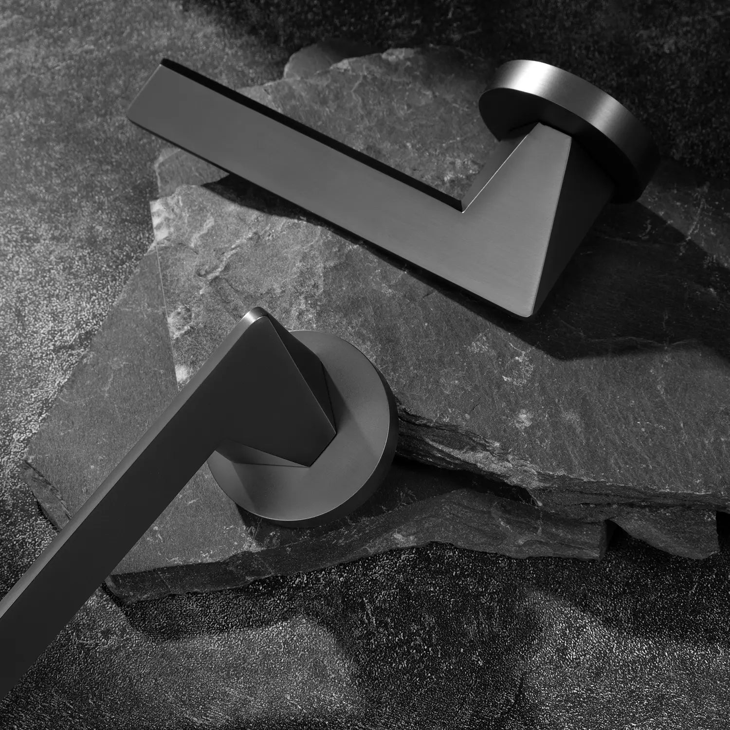 Two Tagg internal door handles in charcoal grey finish, featuring a minimalist geometric lever design and round rose, displayed on two overlapping stone slabs.