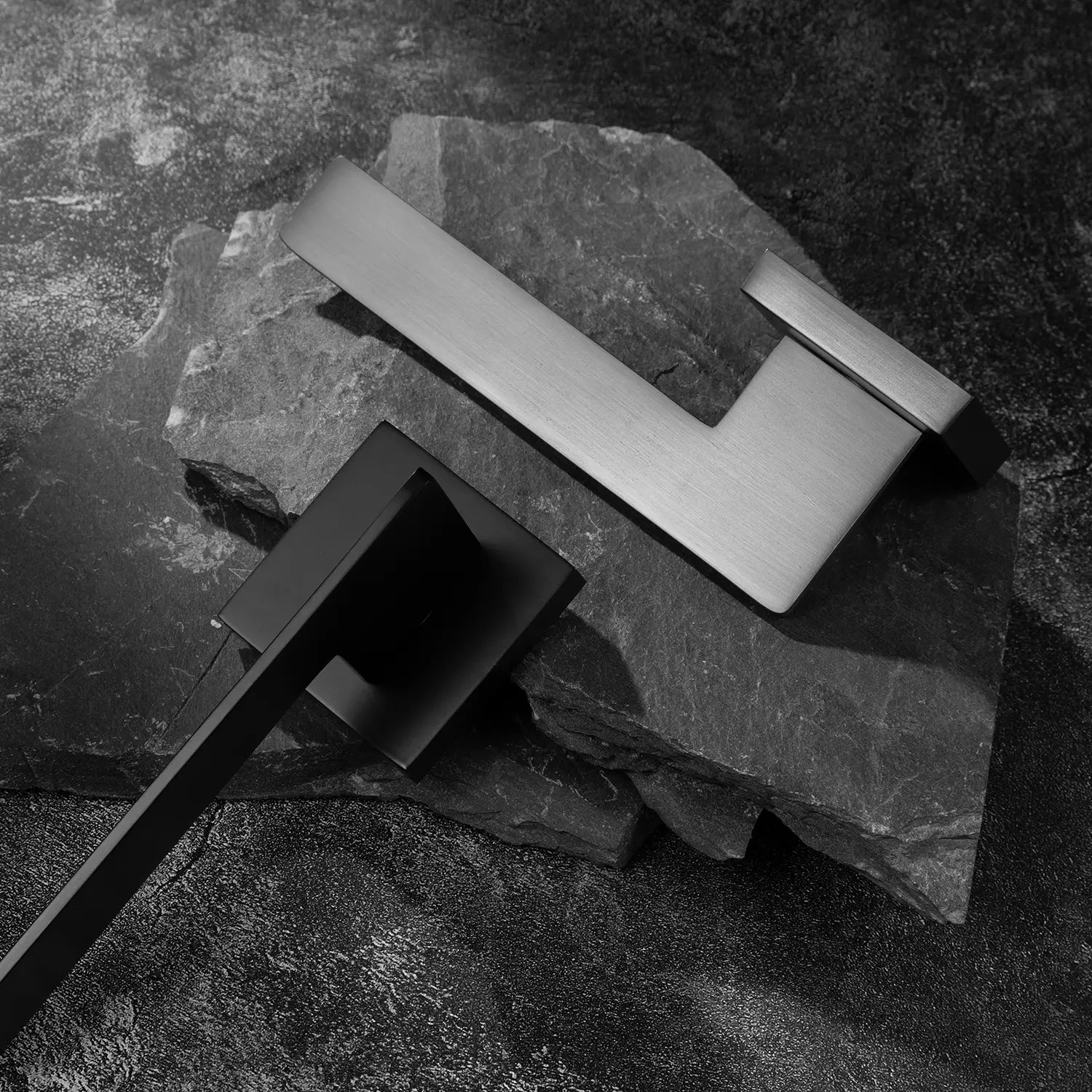 Two Tagg internal door handles in charcoal grey and black finishes, featuring a minimalist geometric lever design and square rose, displayed on two overlapping stone slabs.