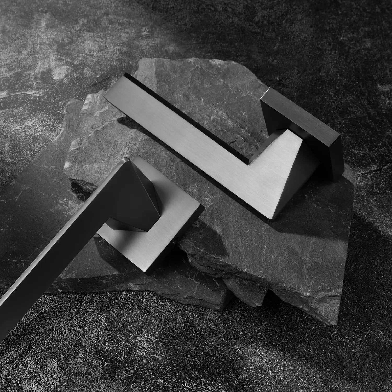 Two Tagg internal door handles in charcoal grey finish, featuring a minimalist geometric lever design and square rose, displayed on two overlapping stone slabs.