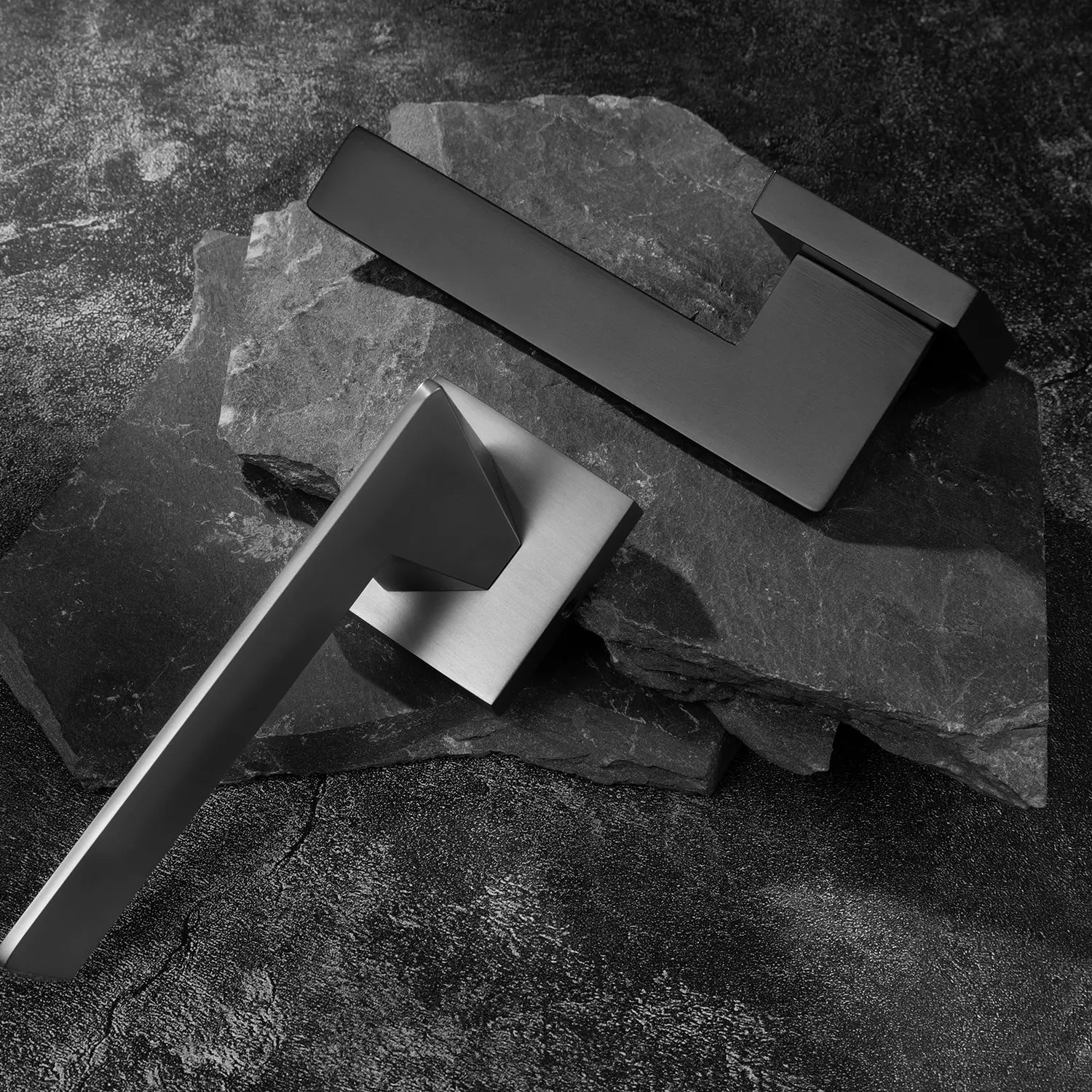 Two Tagg internal door handles in charcoal grey and black finishes, featuring a minimalist geometric lever design and square rose, displayed on two overlapping stone slabs.